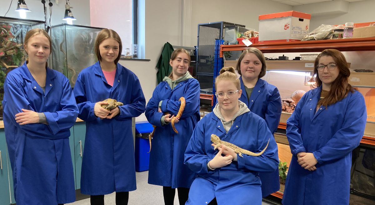 Five students, three holding animals, with lecturer, all wearing blue lab coats in animal house